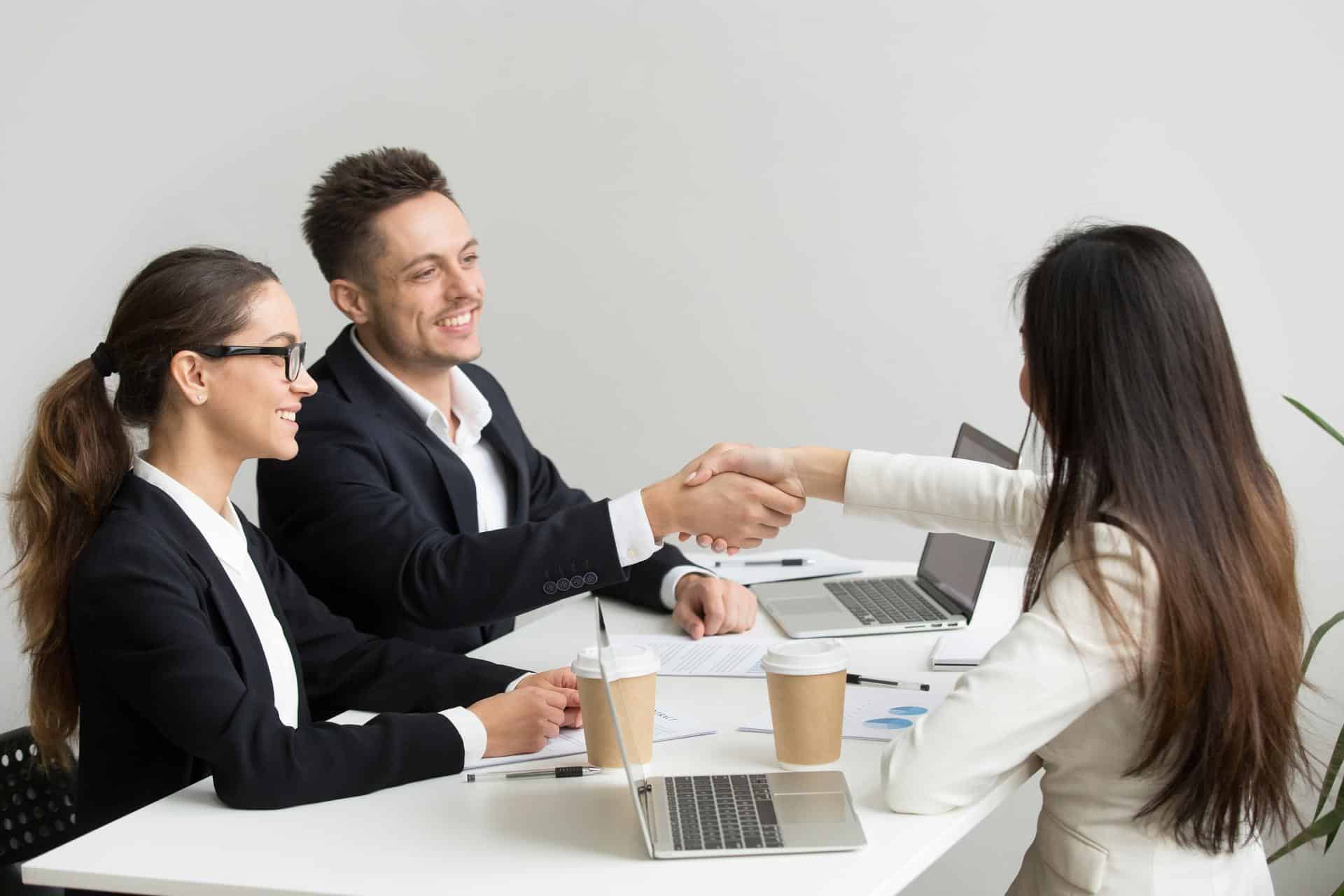 Two smiling recruiters shaking hands with a technical candidate across a desk after an automated technical screening process