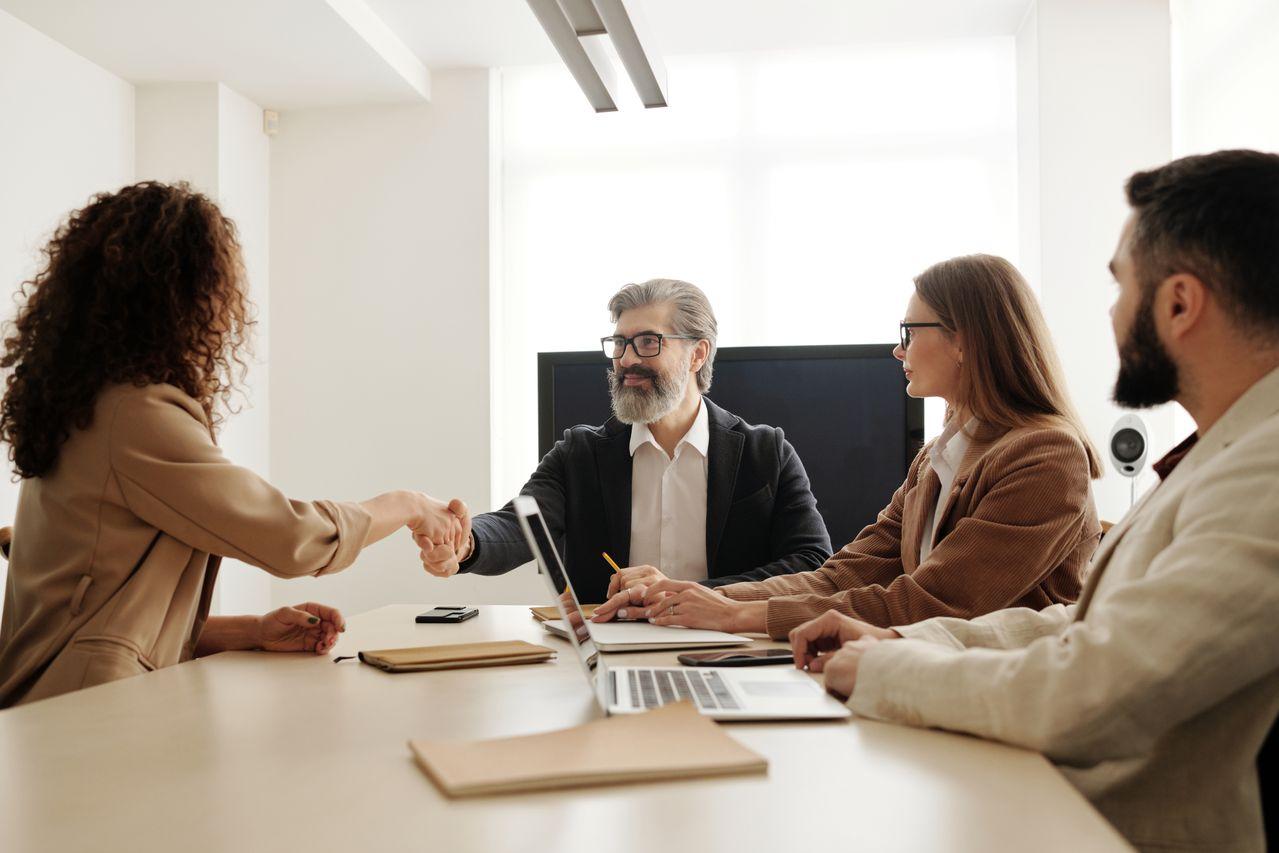 New employee shaking hands with a manager across a conference table while colleagues with laptops look on, starting a structured onboarding process