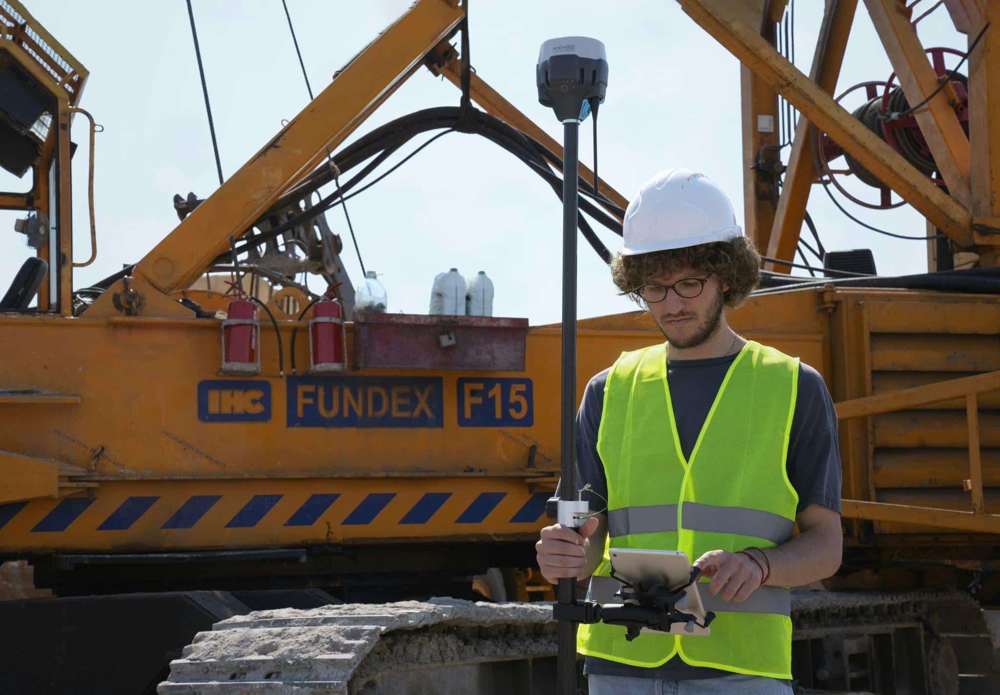 Construction surveyor in a hard hat and safety vest using an RTK GNSS rover and data collector beside a drilling rig, showing which correction approach fits different Canadian projects