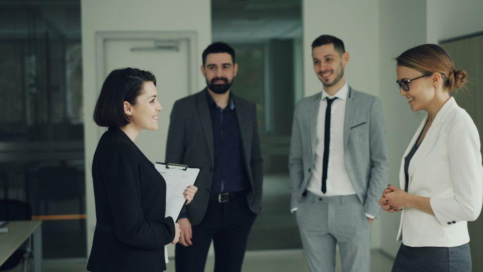 Smiling professional holding a clipboard leads a conversation with three colleagues in a modern office, showing everyday leadership skills