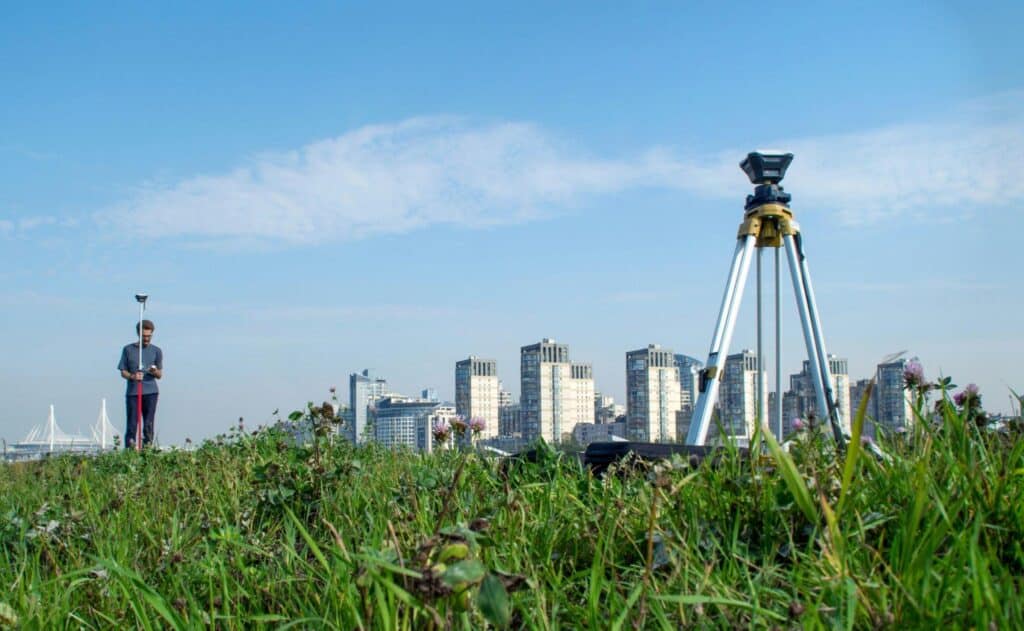 Surveyor holding a GNSS rover pole in a grassy field while a local RTK base station stands on a tripod in the foreground, comparing NTRIP service with local base stations
