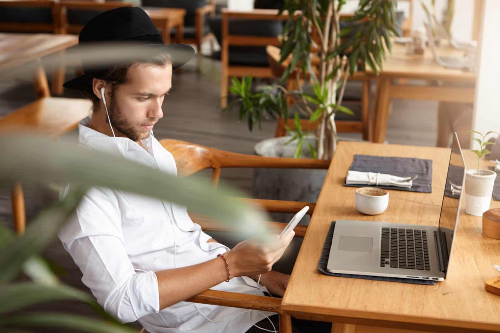 Remote contractor in a hat checking his phone beside an open laptop at a cafe table