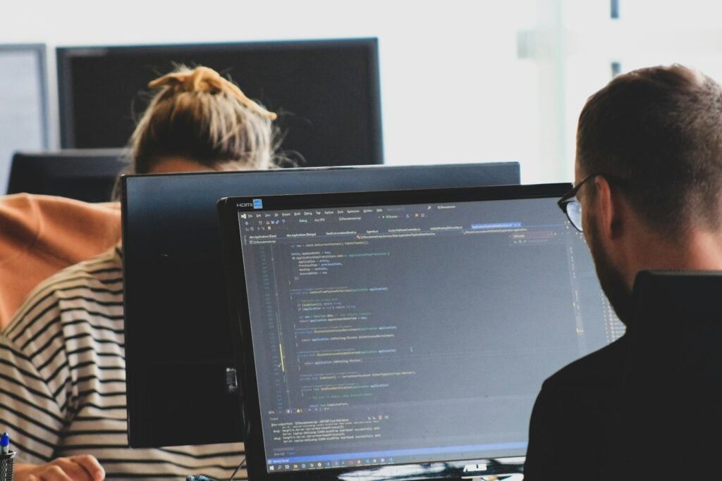 Two security team members working side by side on code at computer monitors in a bright office