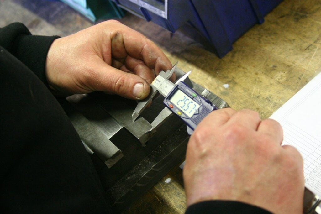 Technician measuring a metal component with a digital caliper beside a technical drawing on a workshop bench
